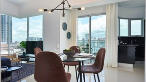 A modern dining area with a glass table, four chairs, a bowl of fruit, and a city view. - Getaway 1BR-WiFi-AC-View-Gym-BlueMall-YellowKey (Santo Domingo)