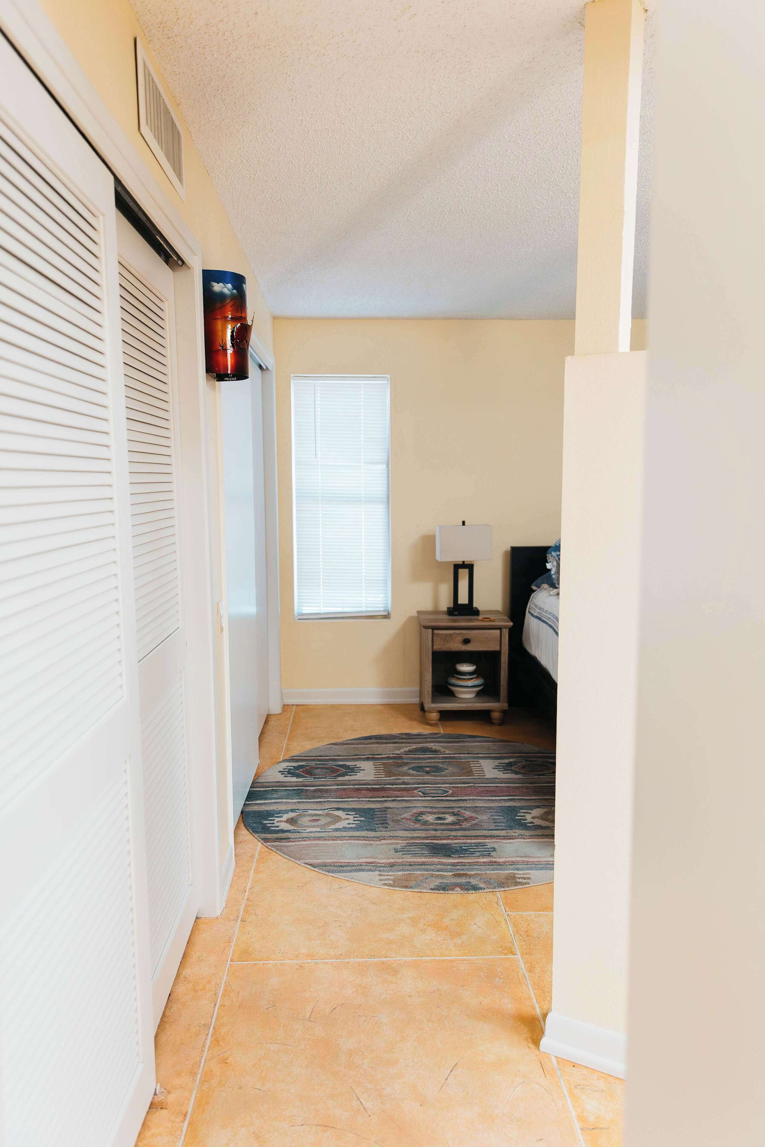 A hallway with a patterned rug, a small table with a lamp, and a window with blinds.
