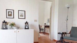 A well-lit living room with a sofa, a dining table, and a lamp. There are framed pictures on the wall and a vase with flowers on the table. - Qanat House_apartment with terrace- In the countryside in the heart of the city (Palermo)