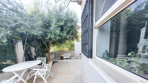 A patio with white furniture, a tiled floor, and a view of a garden with trees and a window. - Qanat House_apartment with terrace- In the countryside in the heart of the city (Palermo)