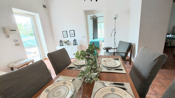 A dining area with a wooden table set for four, surrounded by chairs. There is a view of a balcony through the window, and the room has a modern design with a chandelier. - Qanat House_apartment with terrace- In the countryside in the heart of the city (Palermo)