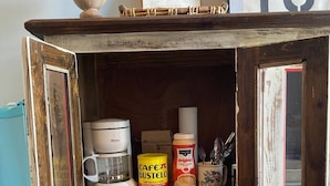 Ein rustikales Holzschränkchen mit Regalen, das eine Kaffeemaschine, eine Kaffeeweißer-Gläser und eine Kaffeepackung sowie ein gerahmtes Bild beherbergt.