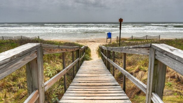 Une promenade en bois mène à une plage. On aperçoit au loin un panneau bleu et un poteau en bois.