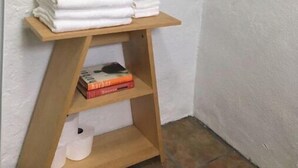 Esquina de una habitación con un estante de madera que sostiene toallas blancas dobladas y una pila de libros.