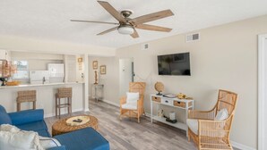 A modern living room with a blue sofa, wicker chairs, a ceiling fan, and a kitchen area in the background.