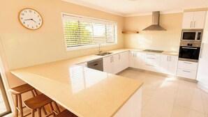 A kitchen with a large island, white cabinets, a stainless steel range hood, and a clock on the wall. - Sea Sounds - Ultimate beachhouse with views (Caves Beach)