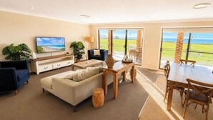 A living room with a sofa, TV, and dining table. - Sea Sounds - Ultimate beachhouse with views (Caves Beach)