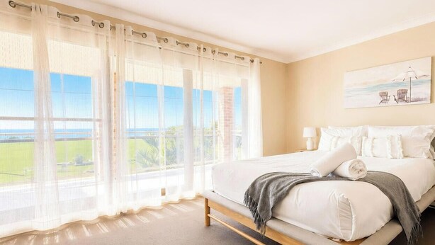 A bedroom with a large window, a bed, a painting, and a view of the outdoors. - Sea Sounds - Ultimate beachhouse with views (Caves Beach)