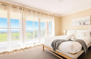 A bedroom with a large window, a bed, a painting, and a view of the outdoors. - Sea Sounds - Ultimate beachhouse with views (Caves Beach)