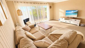 A living room with a sofa, a coffee table, a TV on a stand, and a large window with curtains. - Sea Sounds - Ultimate beachhouse with views (Caves Beach)