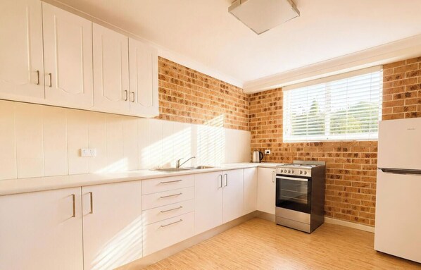 A kitchen with white cabinets, a brick wall, a window, and a built-in oven. - Sea Sounds - Ultimate beachhouse with views (Caves Beach)