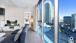 A modern hotel room with a large window offering a city view, a flat-screen TV, a sofa, and a small dining area. - H'Residences – 2 Bedroom Hinterland View — Q Stay (Surfers Paradise)