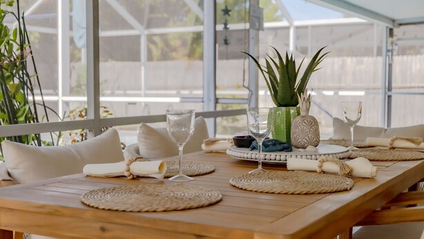Ein Holztisch, gedeckt zum Essen, mit gewebten Platzsets, weißen Kissen und einem Blumentopf.