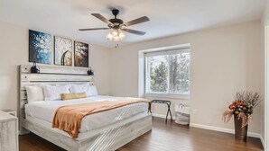 Une chambre à coucher comprenant un lit, un ventilateur de plafond, des œuvres d’art, une fenêtre et un arrangement floral.