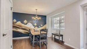 Un coin repas avec une table et des chaises en bois, une fresque murale et une fenêtre avec des stores.