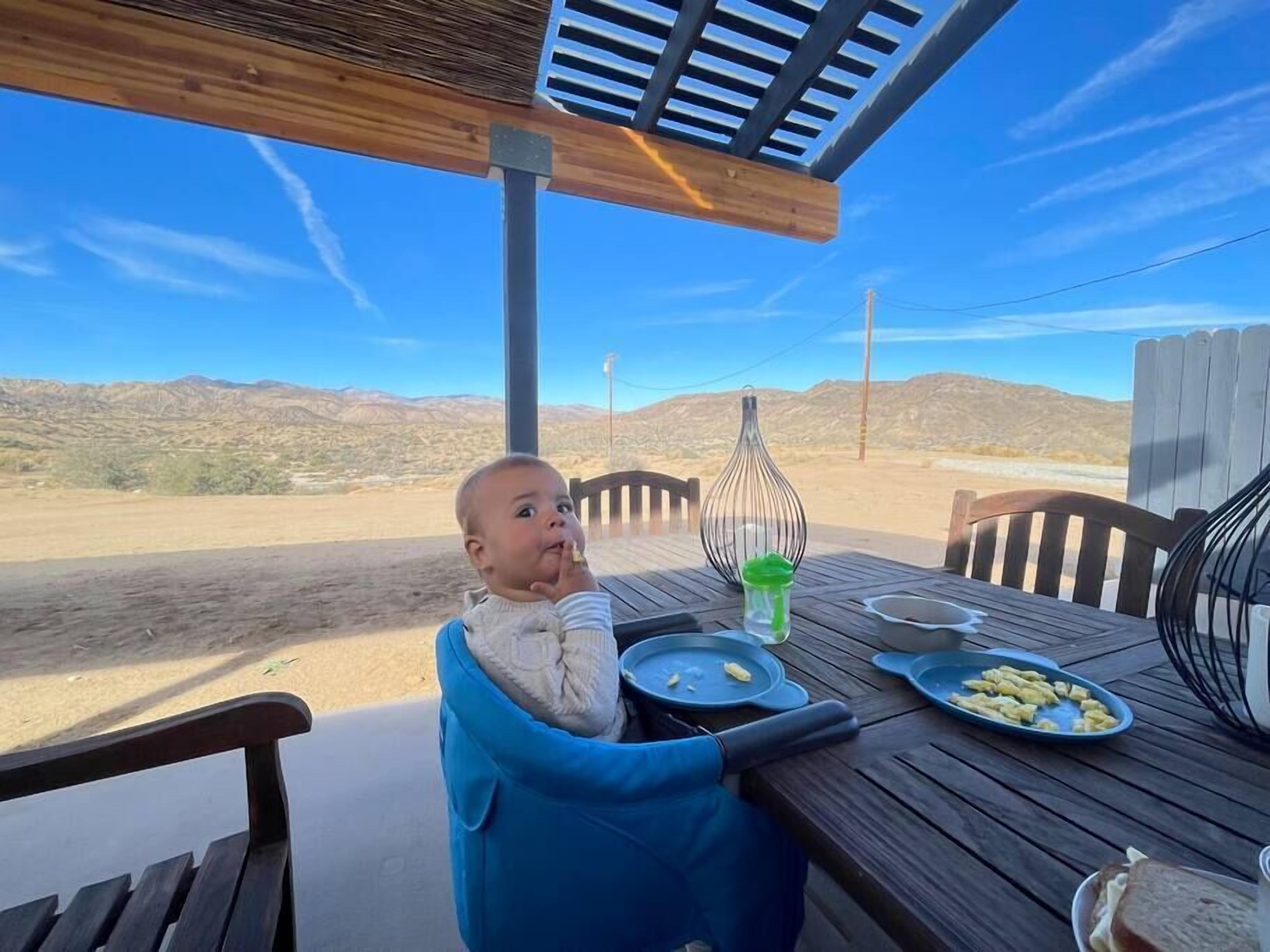 Niño en una trona azul junto a una mesa de madera al aire libre, con platos de comida, una jaula de metal y vistas a un paisaje desértico con montañas al fondo.