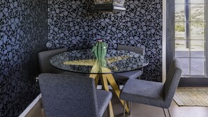 A dining area with a round glass table, four grey upholstered chairs, a green cactus centerpiece, and a wall with a skull pattern. - Chasing The Moon: Walk to Pappy's & Tub Views (Pioneertown)
