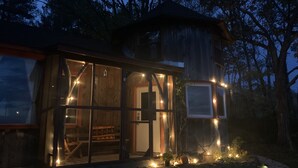 Une cabane en bois rustique, dotée de grandes fenêtres et illuminée de guirlandes lumineuses, et entourée d’arbres.