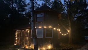 Un chalet rustique doté de grandes fenêtres et d’un porche, entouré d’arbres et illuminé par des guirlandes lumineuses.