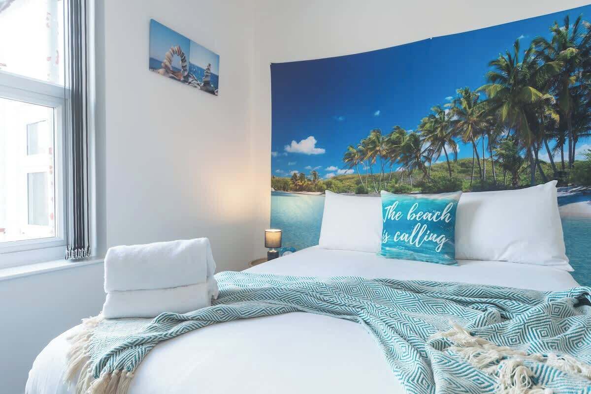 Ein Schlafzimmer mit Bett, Kissen, Decke, Nachttisch, Lampe, Fenster und einer Wand mit einer Stranddarstellung.