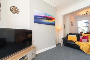 A living room with a flat-screen TV, a modern abstract painting, a black leather sofa with yellow and red cushions, and a window with natural light. - WGcDDE Large 3 Bedroom House - Parking - Garden (Kingston upon Hull)