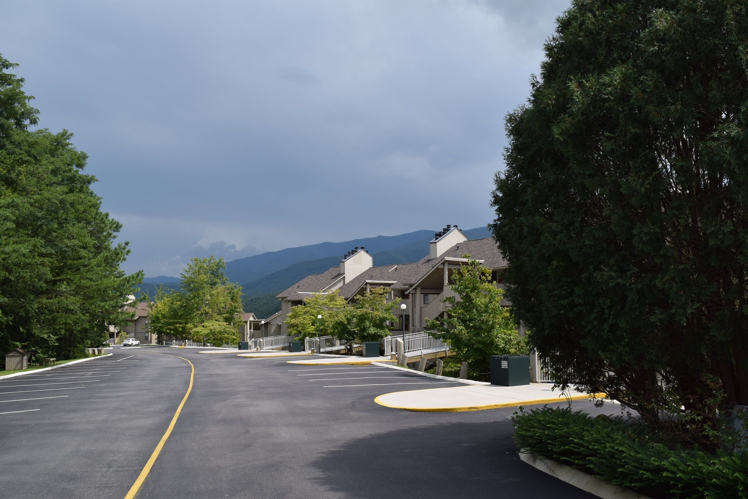 Un parking délimité par une ligne jaune incurvée, entouré d’arbres et de bâtiments, avec une chaîne de montagnes en arrière-plan.