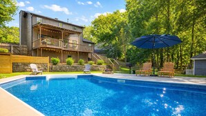 En swimmingpool med ett trädäck och solstolar, omgiven av ett hus med en balkong och en mur av sten.