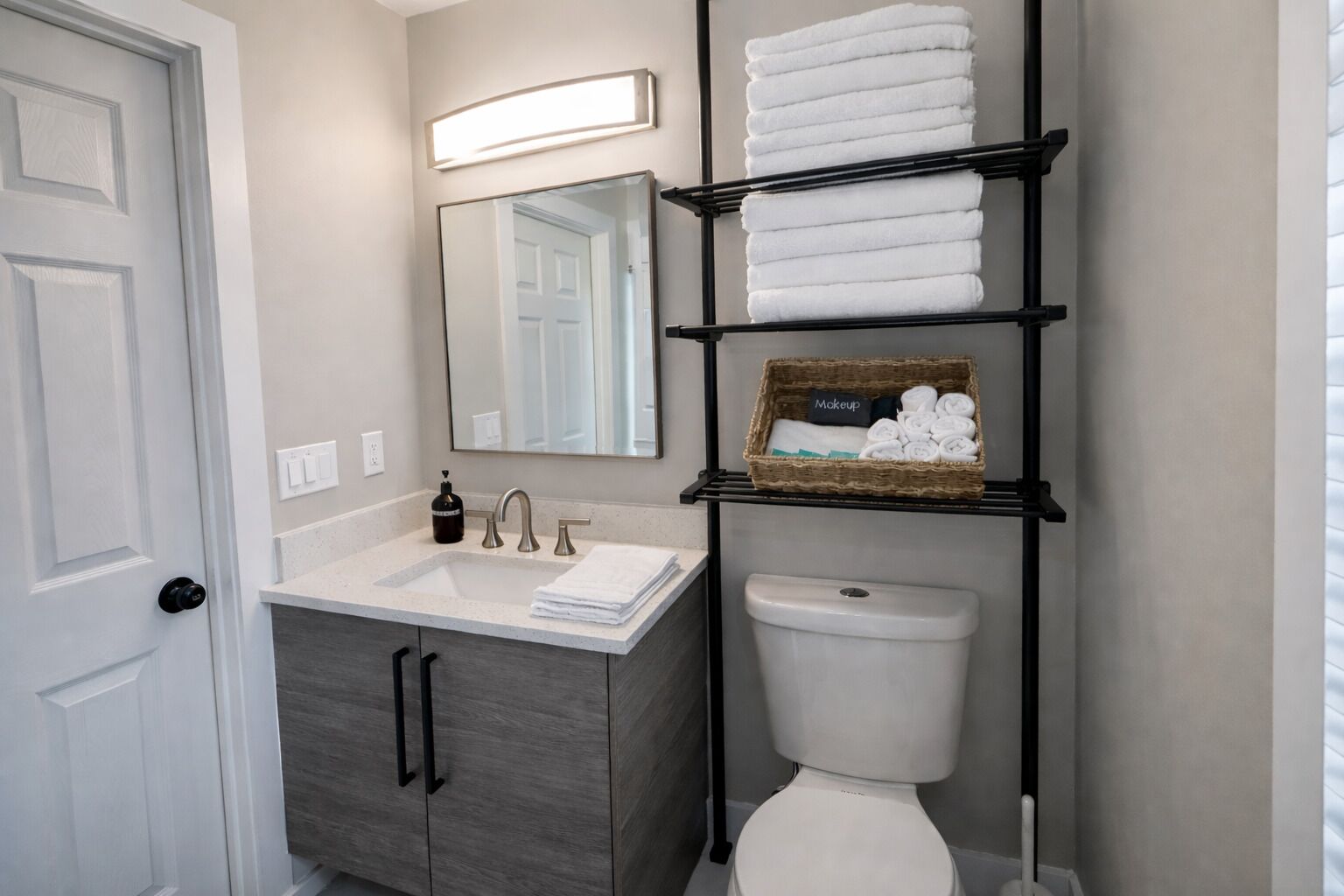 Une salle de bain avec un lavabo blanc, un miroir, des toilettes et des étagères où sont rangées des serviettes et un panier étiqueté « maquillage ».