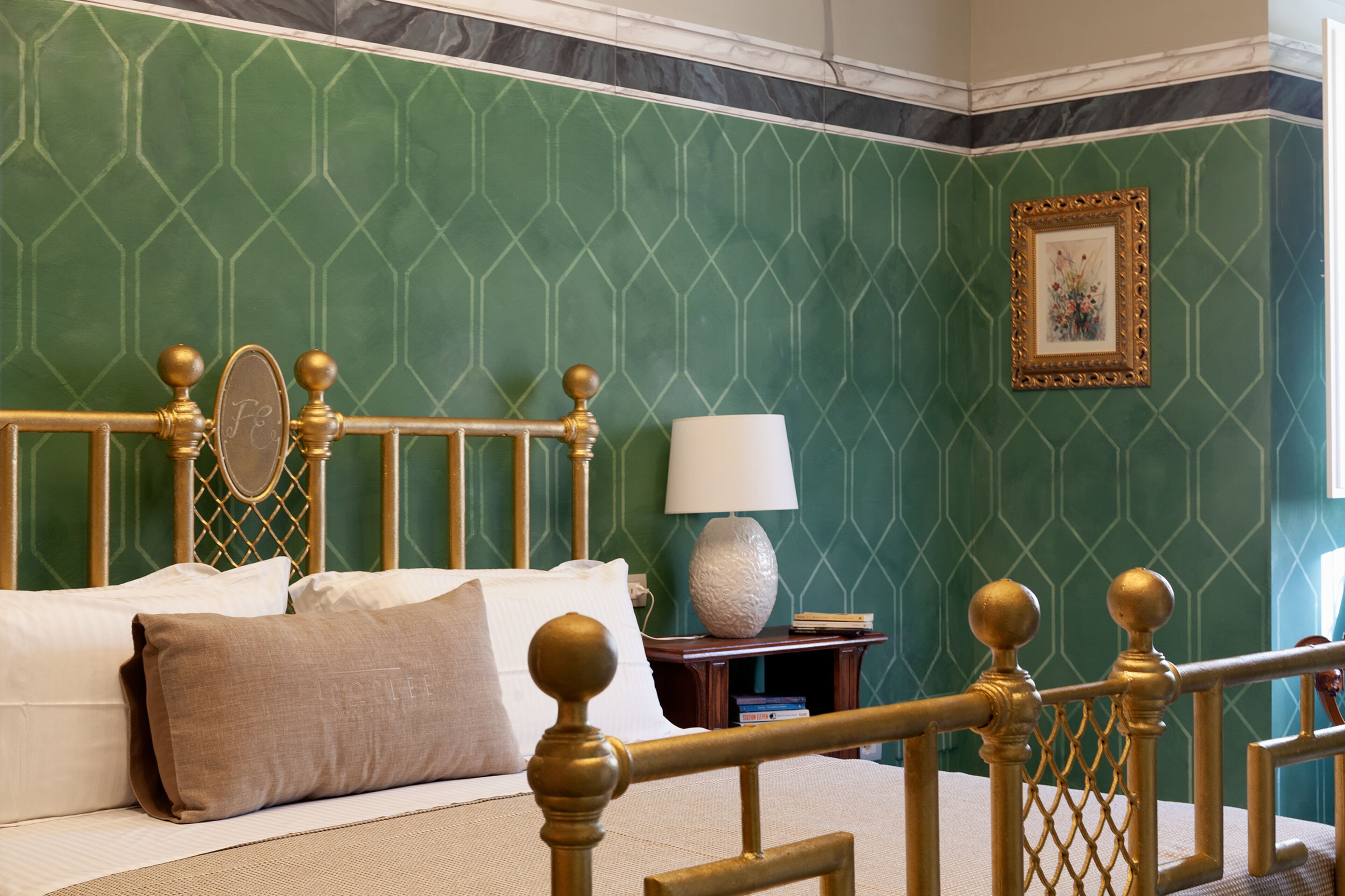 A neatly made bed with a gold headboard, a bedside table with a lamp, and a framed picture on the wall.