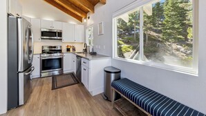 Cozinha compacta com eletrodomésticos modernos, janela com vista para uma floresta e um banco junto à janela.