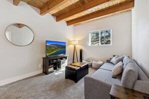 A living room with a flat-screen TV, a sofa, a coffee table, a lamp, and a window with a view of trees.