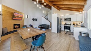 Um interior moderno com mesa de jantar de madeira, cadeiras azuis, lareira, sofá e cozinha com eletrodomésticos de aço inoxidável.
