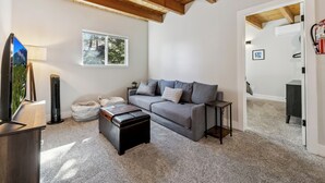 Uma sala de estar moderna com um sofá cinzento, uma pequena mesa lateral, uma televisão de tela plana e teto com vigas de madeira aparente.