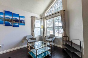 Una habitación con una mesa de vidrio, dos sillas, una estantería y una ventana con cortinas.