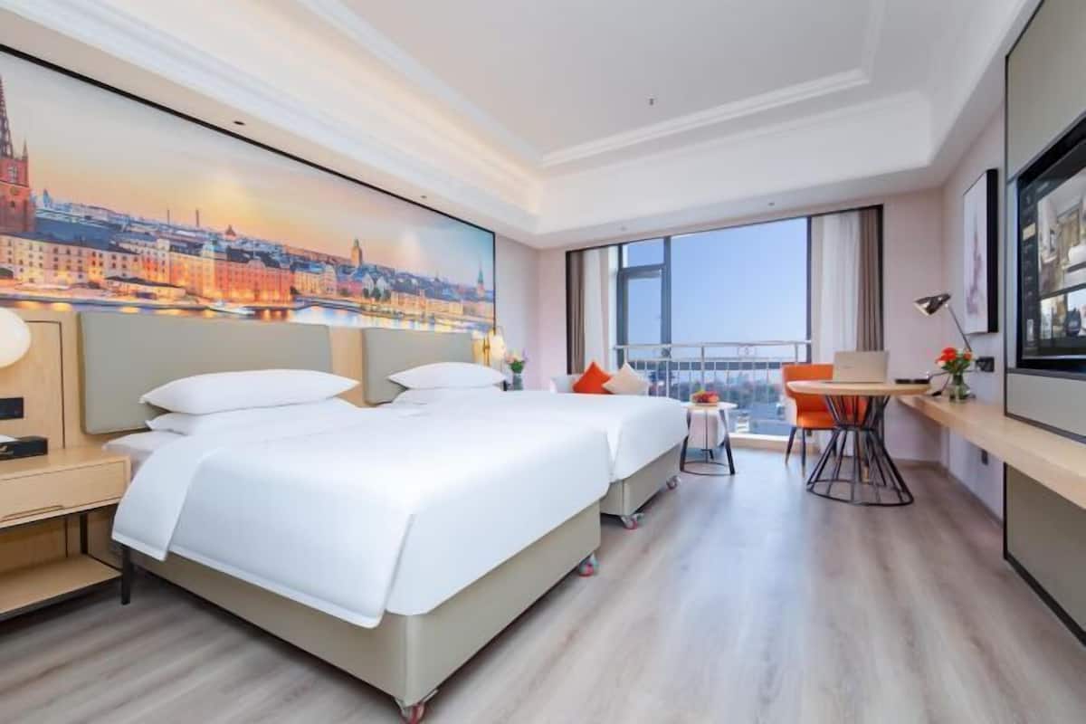 Une chambre d’hôtel avec deux lits, un bureau, une chaise, une fenêtre donnant sur la ville et un tableau au mur.