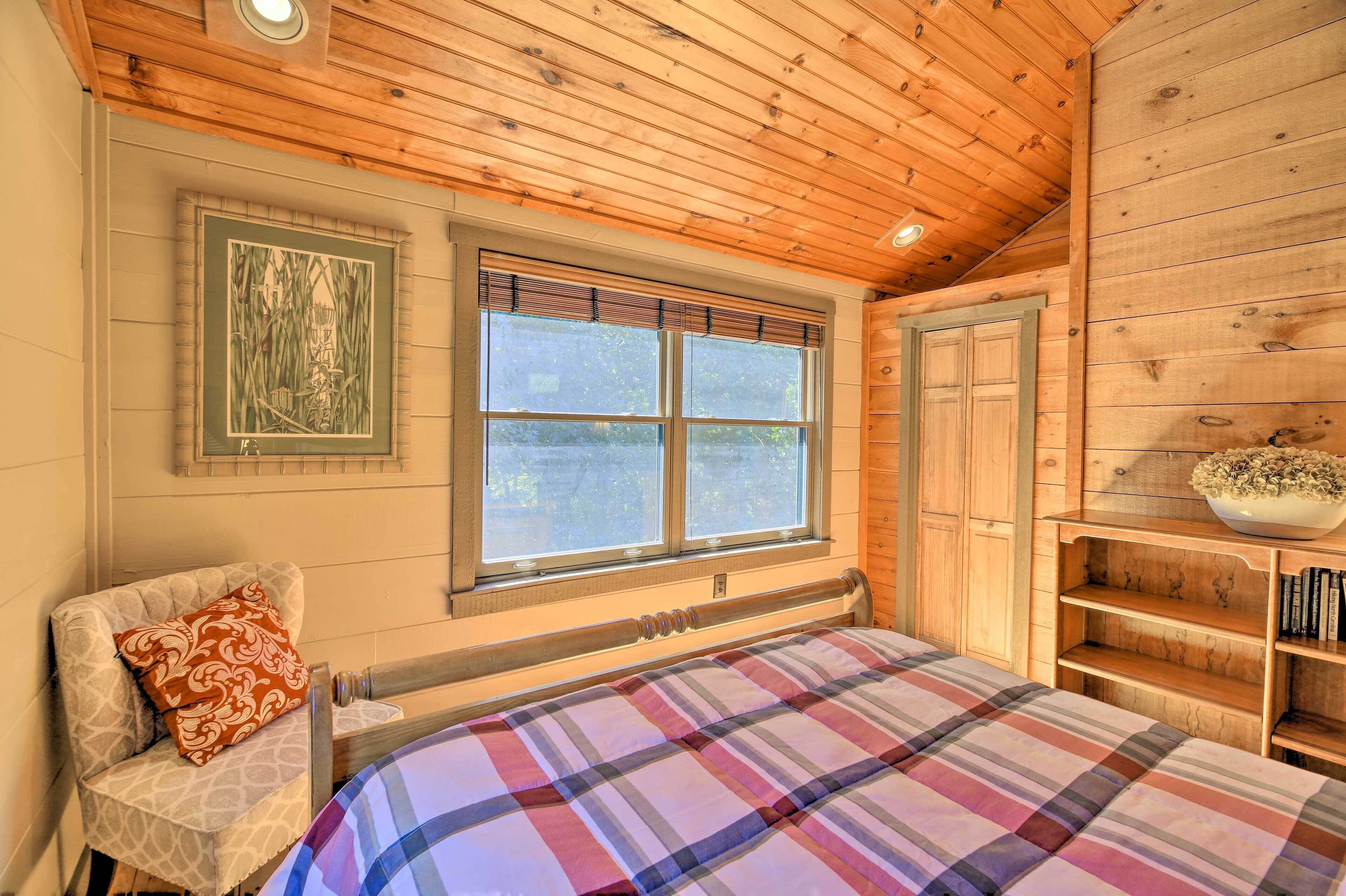 Un dormitorio de cabaña de madera con una cama, una silla, una ventana con persianas y una estantería con libros.