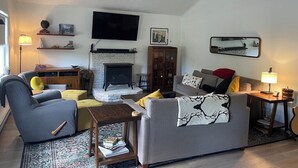 A living room with a fireplace, sofa, armchair, and a television mounted on the wall. - Cozy year round retreat! Close to skiing, hiking and swimming. (East Jewett)