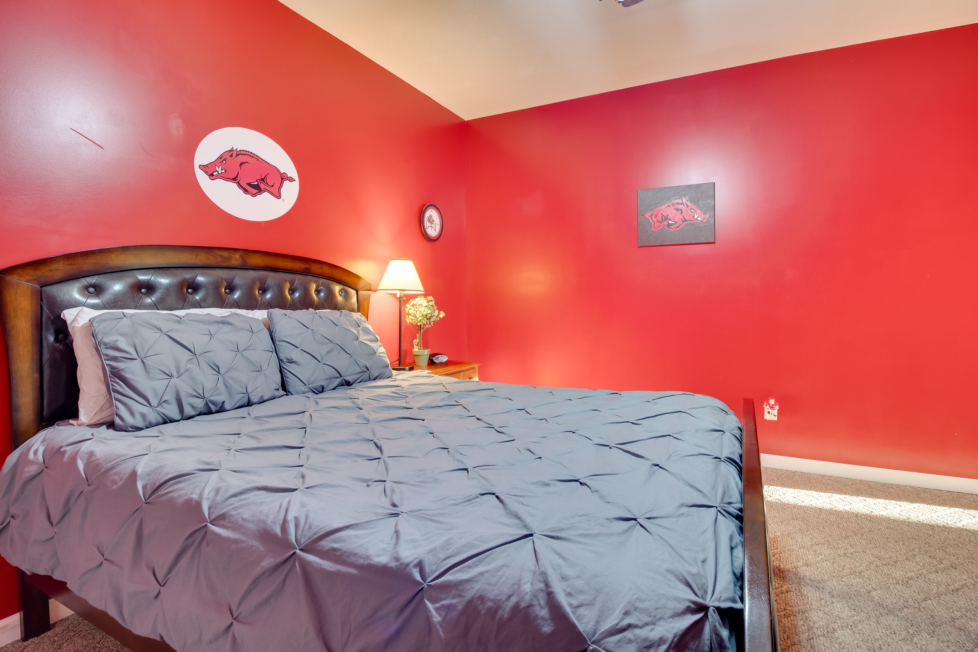A bedroom with a red wall, a bed with a grey quilt, a wooden headboard, a nightstand with a lamp, and a wall clock.