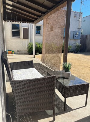 An outdoor patio area with wicker furniture and a glass table, a covered seating area, and a building with windows and a fence in the background.