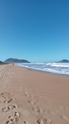 A sandy beach with footprints leading towards the ocean, a clear sky, and distant hills. - Your home in Floripa! (SC)