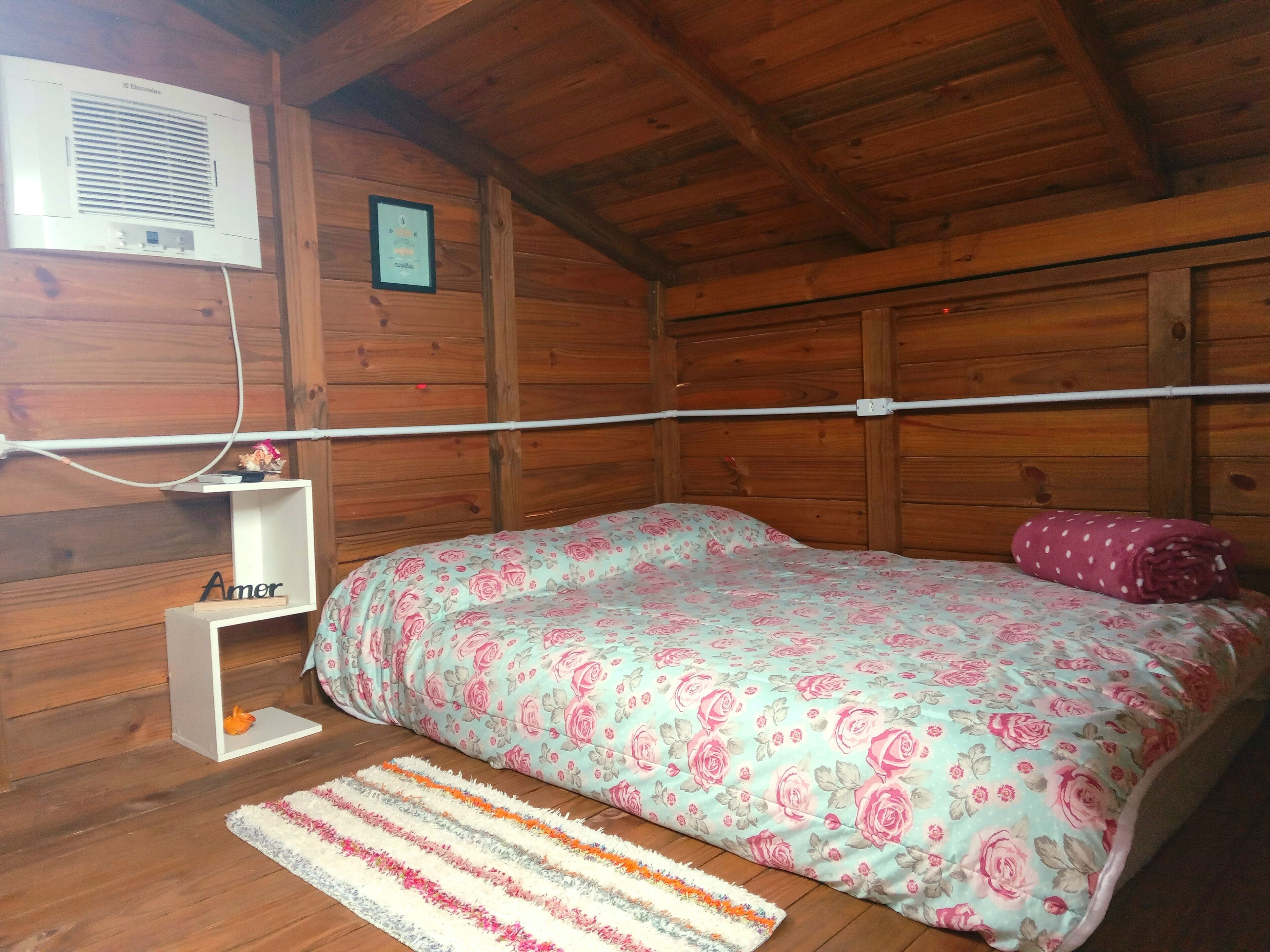 Een houten slaapkamer met een bed, een klein plankje met naamplaatje, een lijst met afbeelding en een airconditioning.