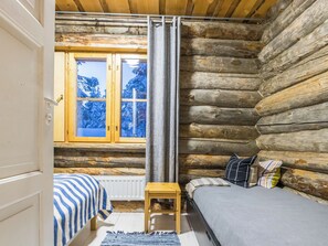 Une chambre rustique en rondins de bois, avec un lit, un petit tabouret et une fenêtre donnant sur des arbres couverts de neige.