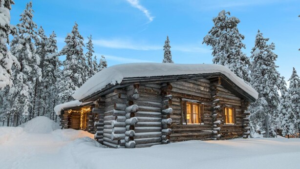 A log cabin with a snow-covered roof and windows, surrounded by snow-laden trees.