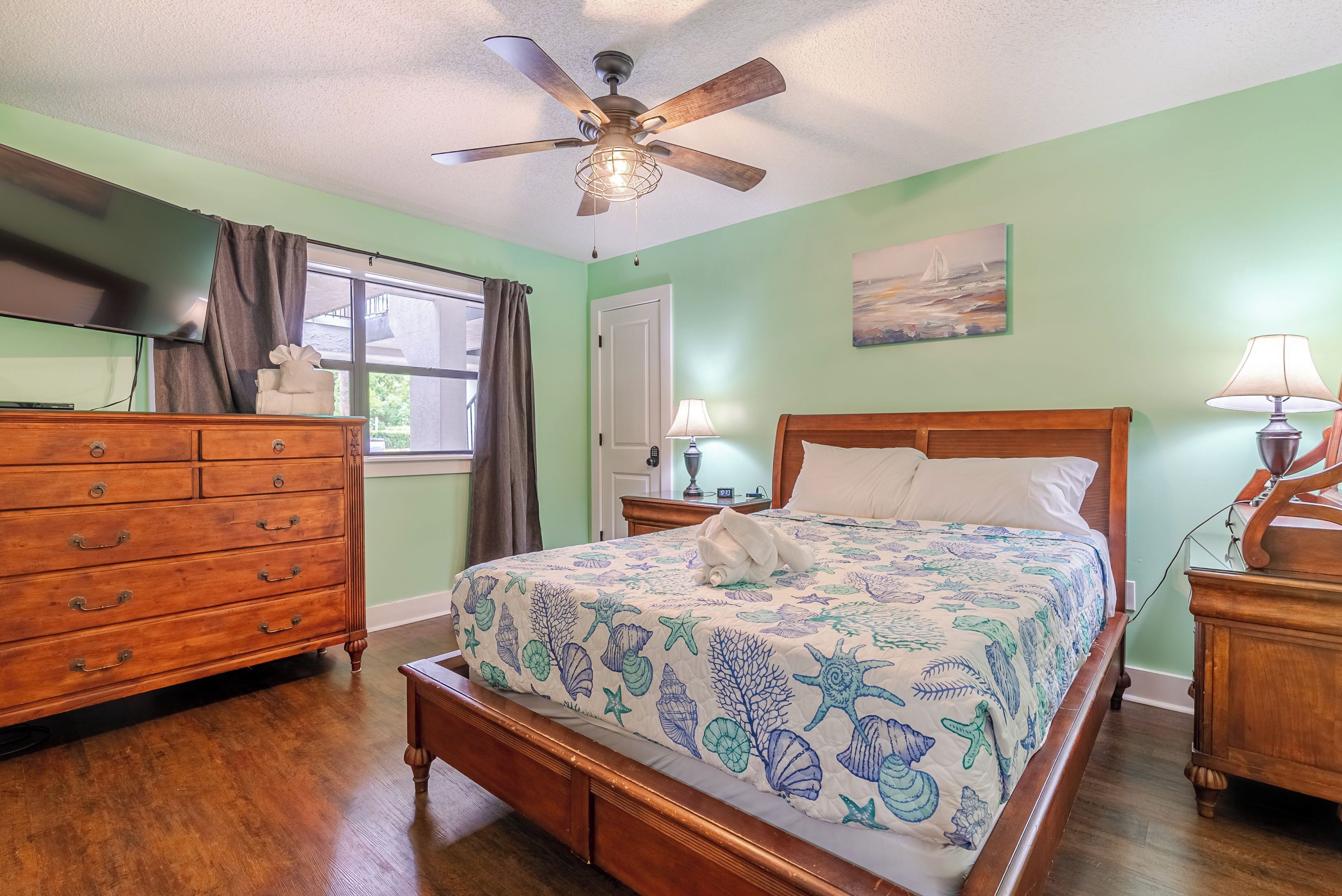 A bedroom with a bed, dresser, ceiling fan, and a window with curtains.