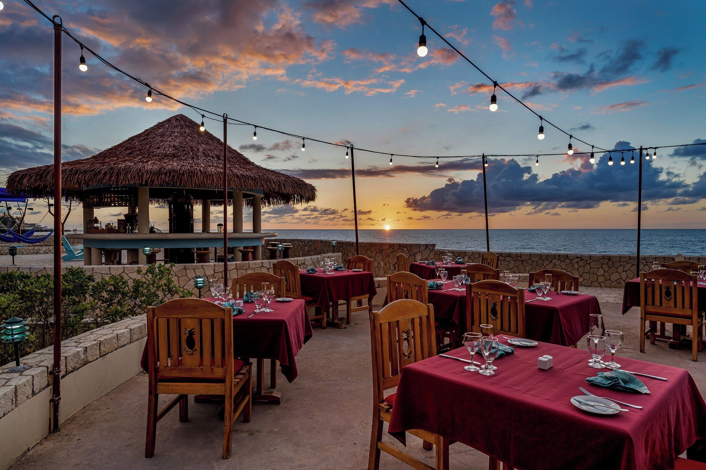 An outdoor dining area with tables and chairs, a thatched-roof structure, and a view of the sunset over the ocean.