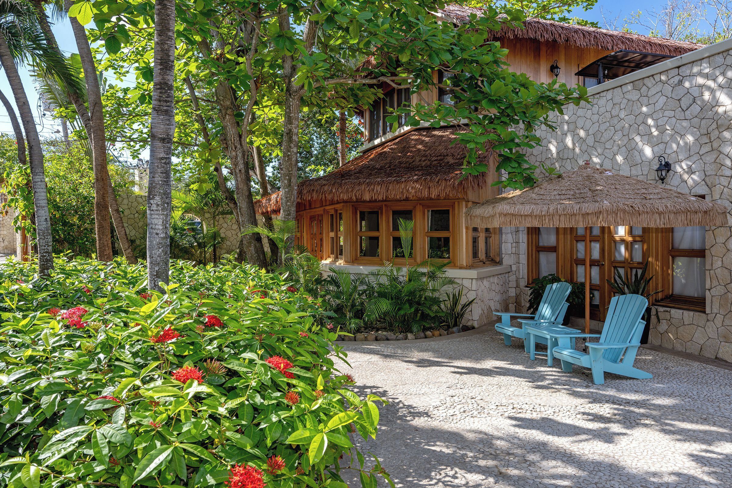 A tropical-style building with a thatched roof, stone walls, and a gravel patio with blue chairs.