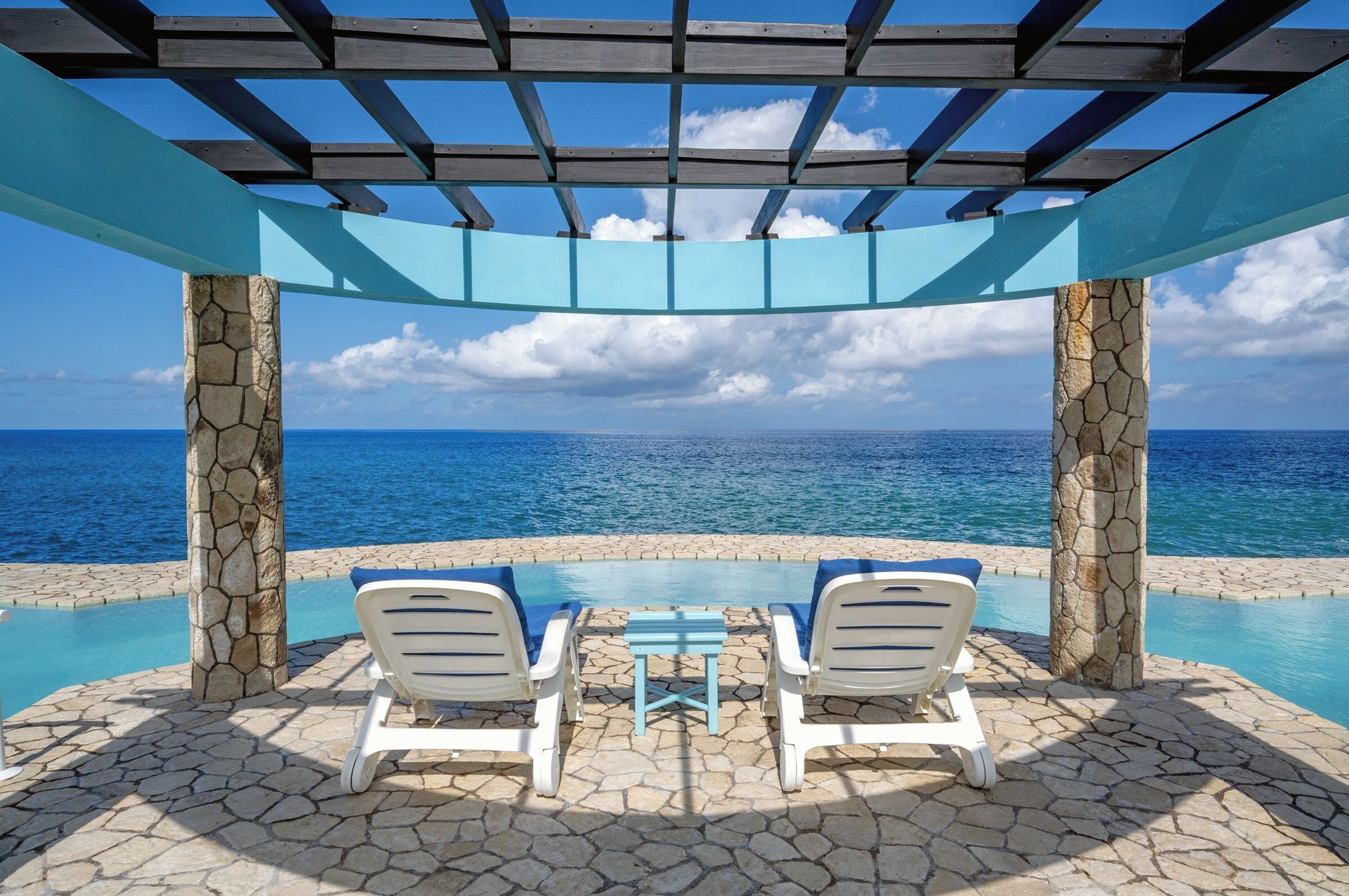 A poolside area with two lounge chairs and a table, overlooking a clear blue sea under a wooden pergola.