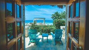 Une vue depuis une fenêtre à cadre en bois donnant sur un patio avec deux chaises Adirondack bleues, une petite table et un mur de pierre orné de fleurs rouges.