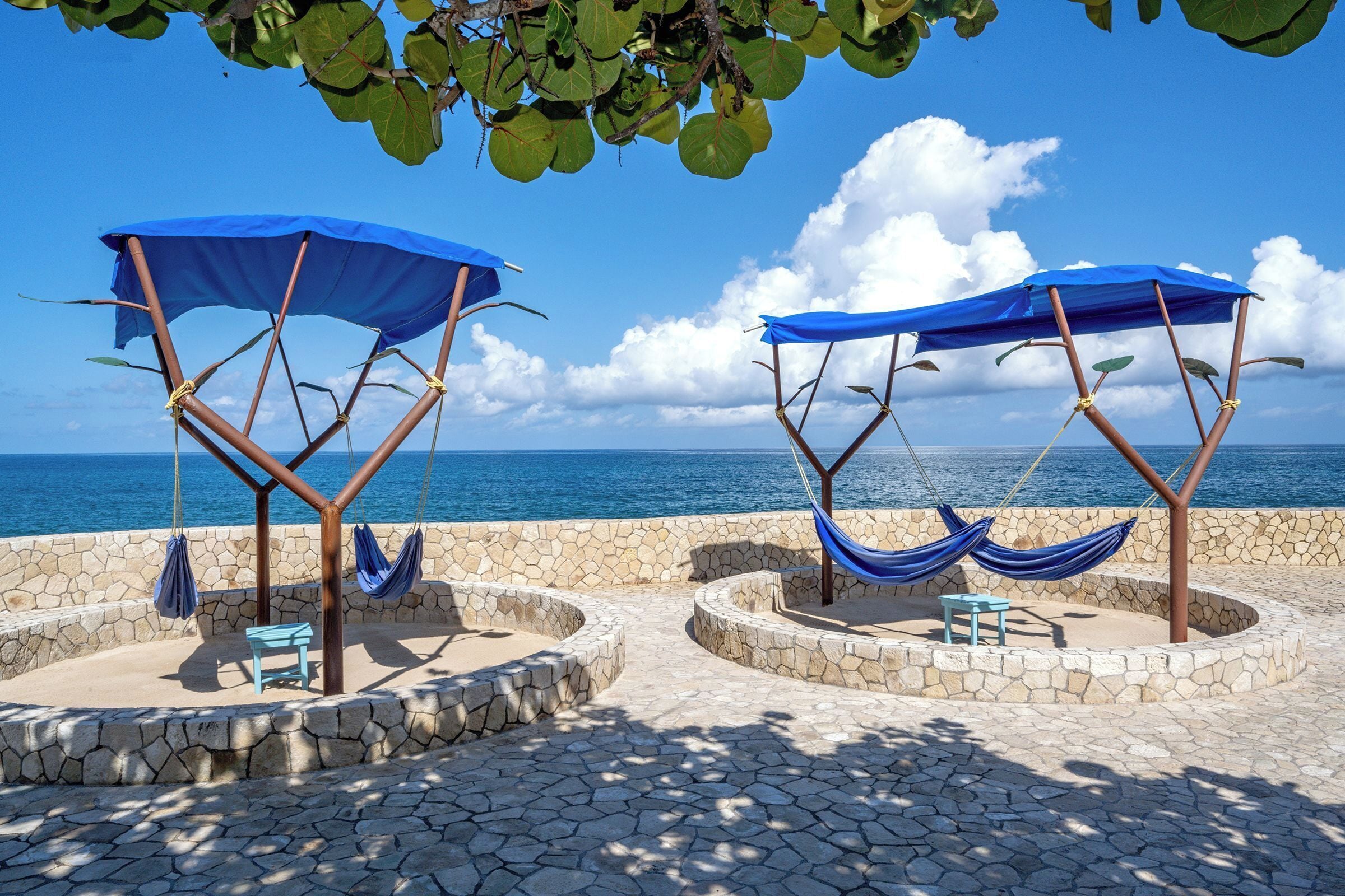 Two hammocks under blue canopies, a stone wall, and a view of the sea.