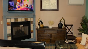 A living room with a fireplace, a television mounted above it, a wooden cabinet, and a glass coffee table. - Comfortable private unit with hot tub, swimming pool and free parking (Gibsonia)